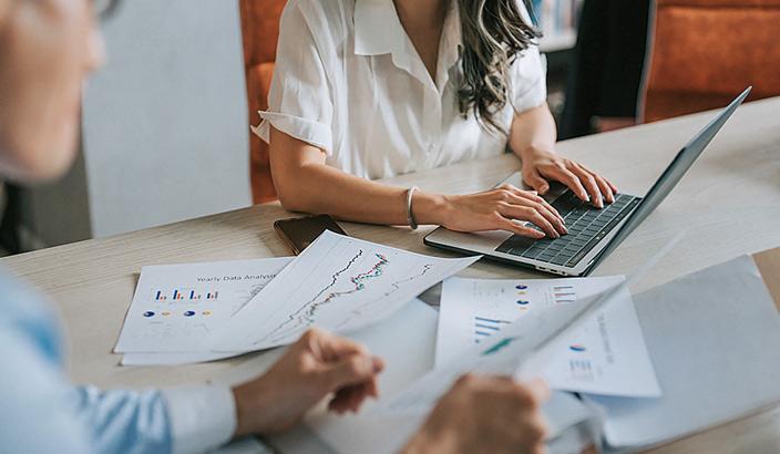 Une entrepreneure qui consulte des documents contenant des graphiques accompagnée d'une autre entrepreneure devant son ordinateur 